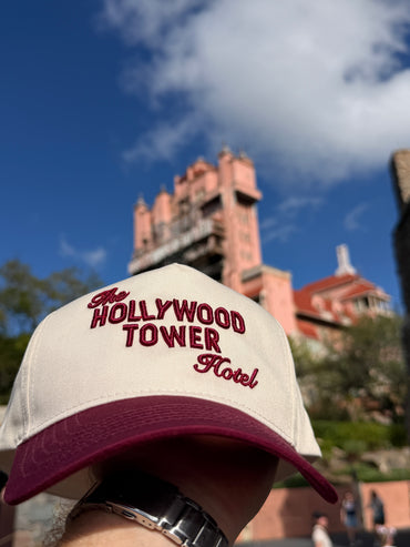 Cream and burgundy Hollywood Tower Hotel hat held in front of pink themed building under blue sky