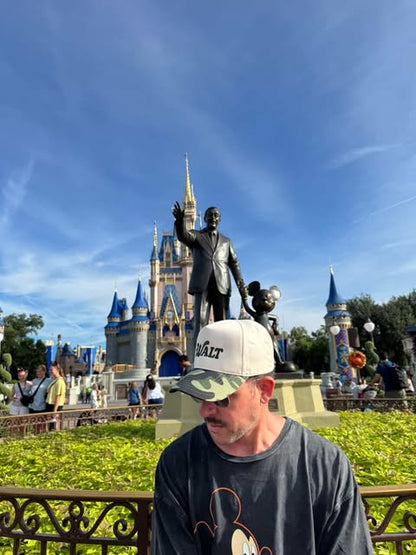 Man in Mickey Mouse shirt and Walt hat at Cinderella Castle, Disney park, sunny day