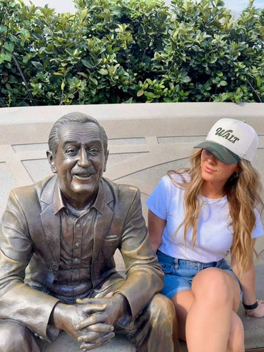 Woman in white shirt and Walt cap sitting beside bronze statue on outdoor bench