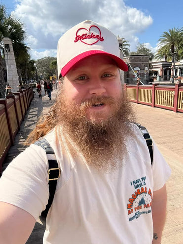 Man in Mickey heart hat and Disney shirt smiling outdoors at theme park entrance