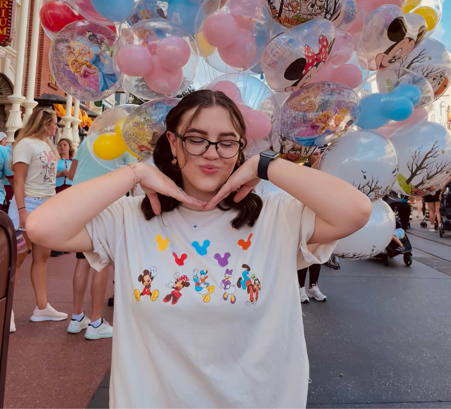 Person holding a large bouquet of balloons with cartoon characters, wearing a white t-shirt with colorful graphics, in an outdoor setting.