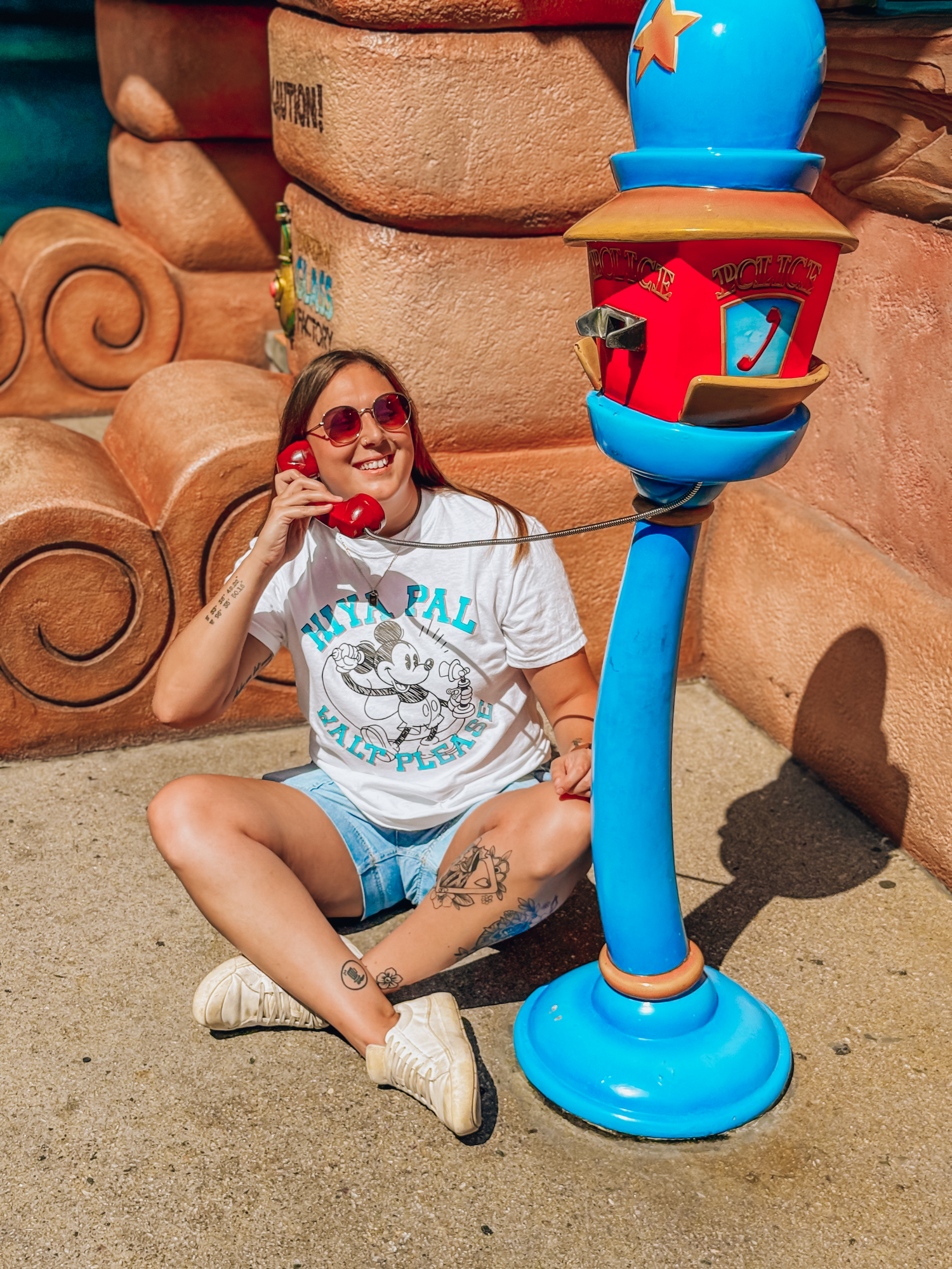 Woman wearing Disney-inspired cartoon t-shirt and sunglasses, sitting on sidewalk, smiling with red phone prop at a colorful theme park.