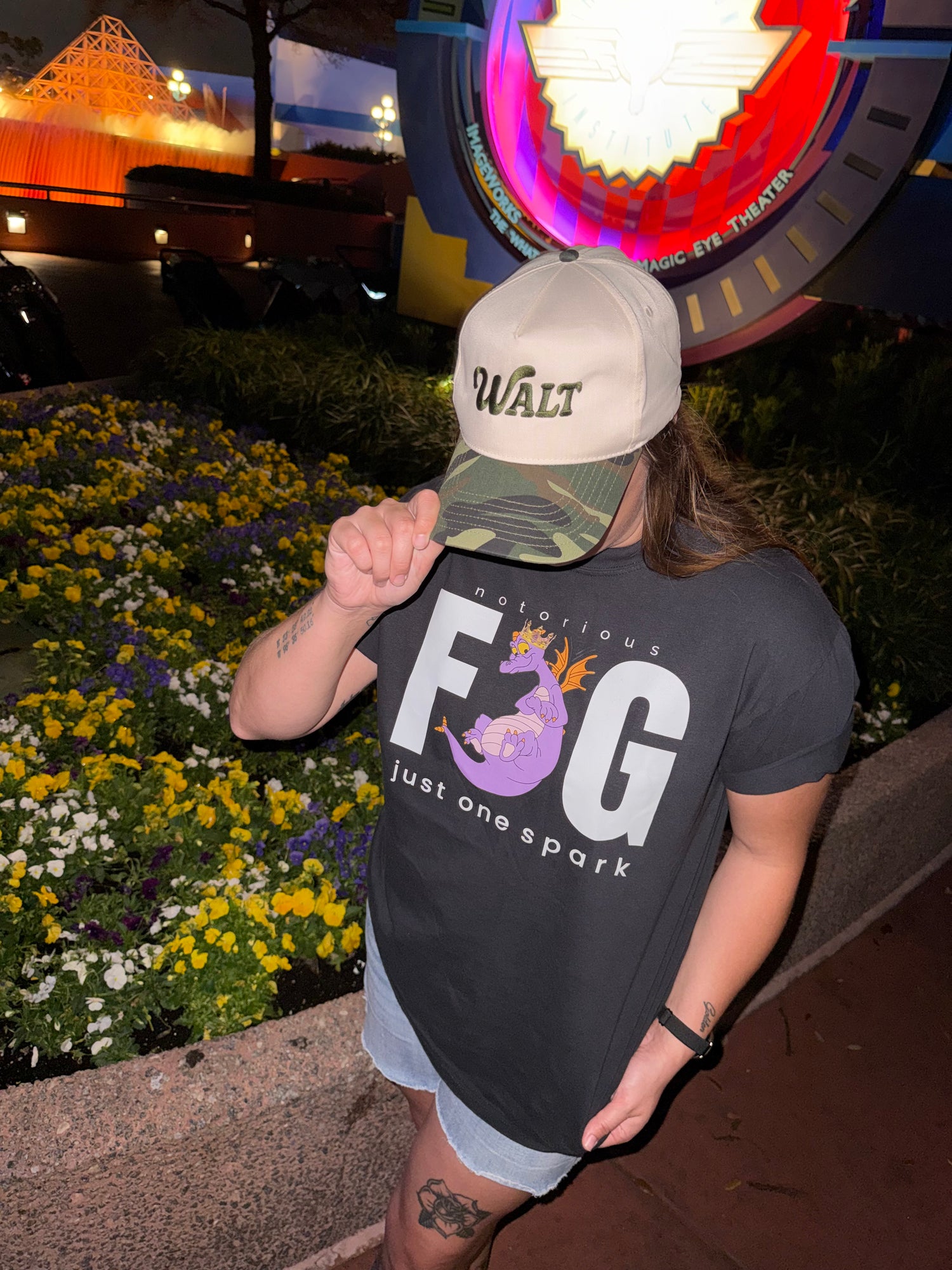 Person in a Walt hat and Figment graphic t-shirt at Epcot, standing near flowers at night.