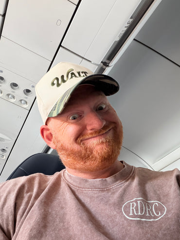 Smiling man with red beard in 'WALT' cap and RDRC shirt on airplane seat