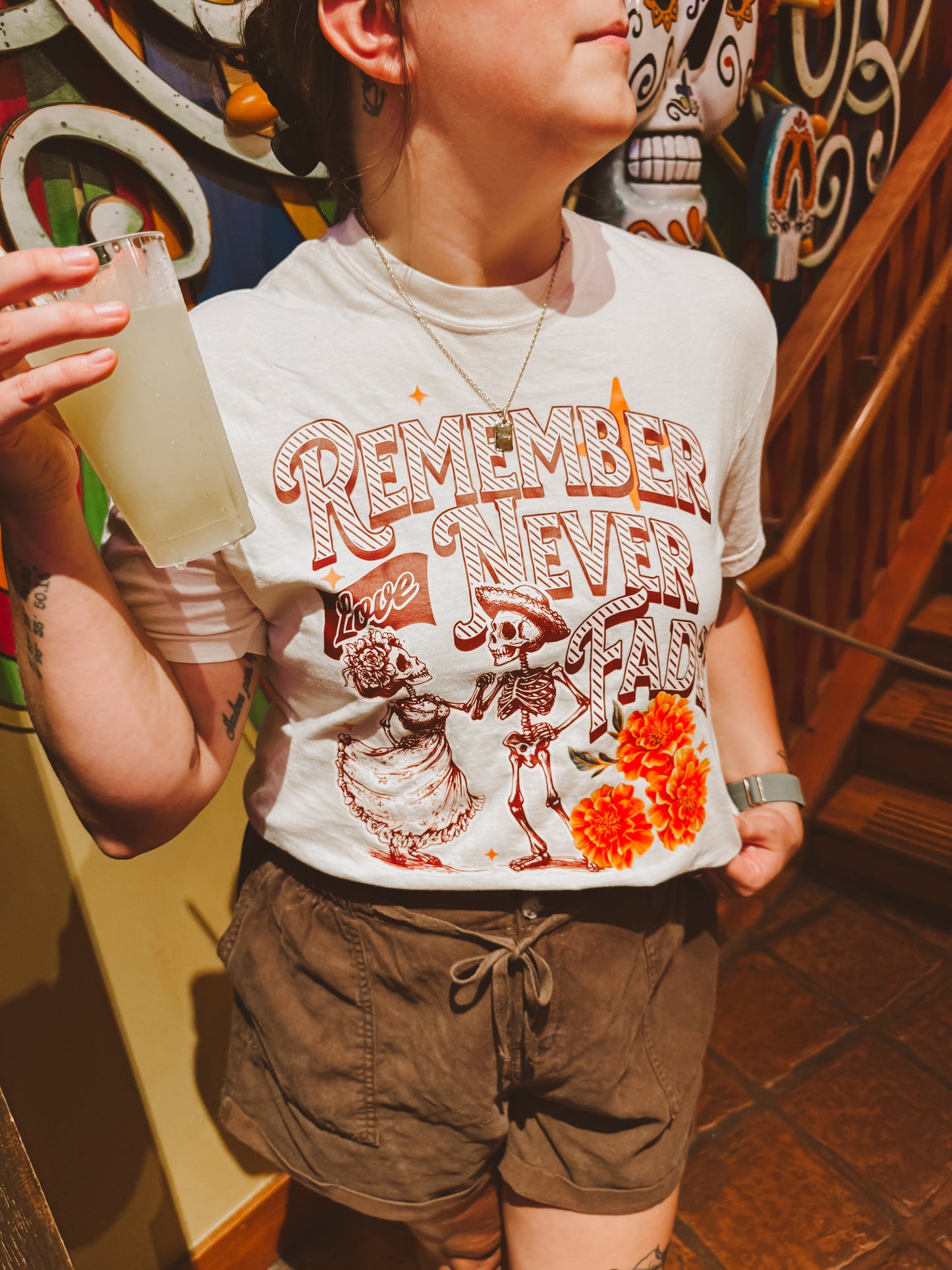Person wearing Disney-inspired Day of the Dead shirt with skeletons and marigolds, holding a drink