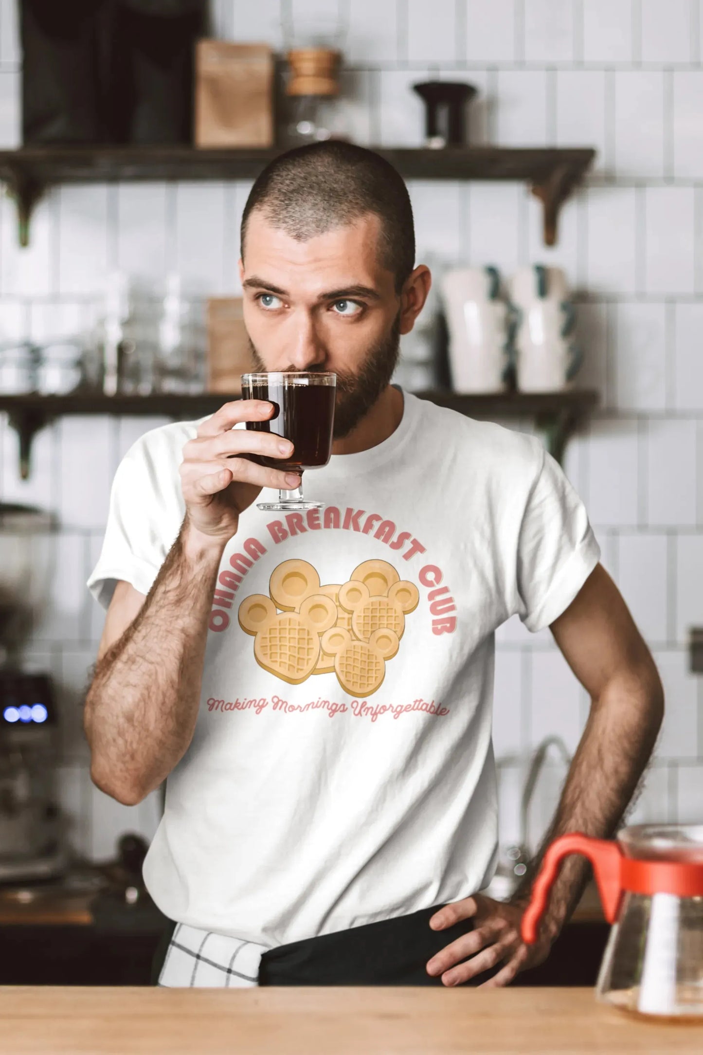 Man in kitchen wearing white Ohana Breakfast Club shirt with Disney-style waffles, drinking coffee