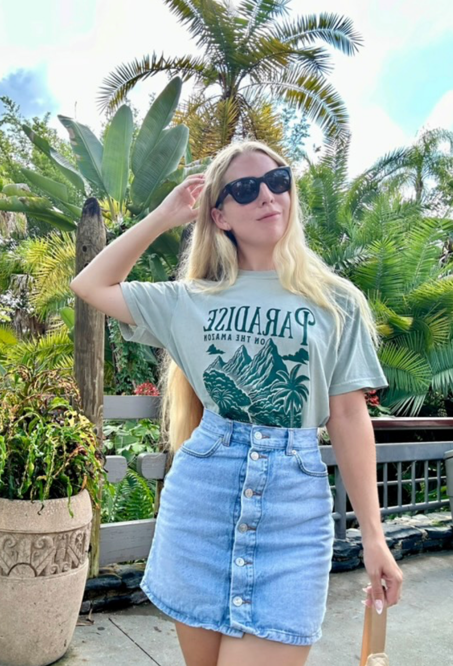 Woman in sunglasses wearing a Paradise Amazon graphic t-shirt and denim skirt outdoors