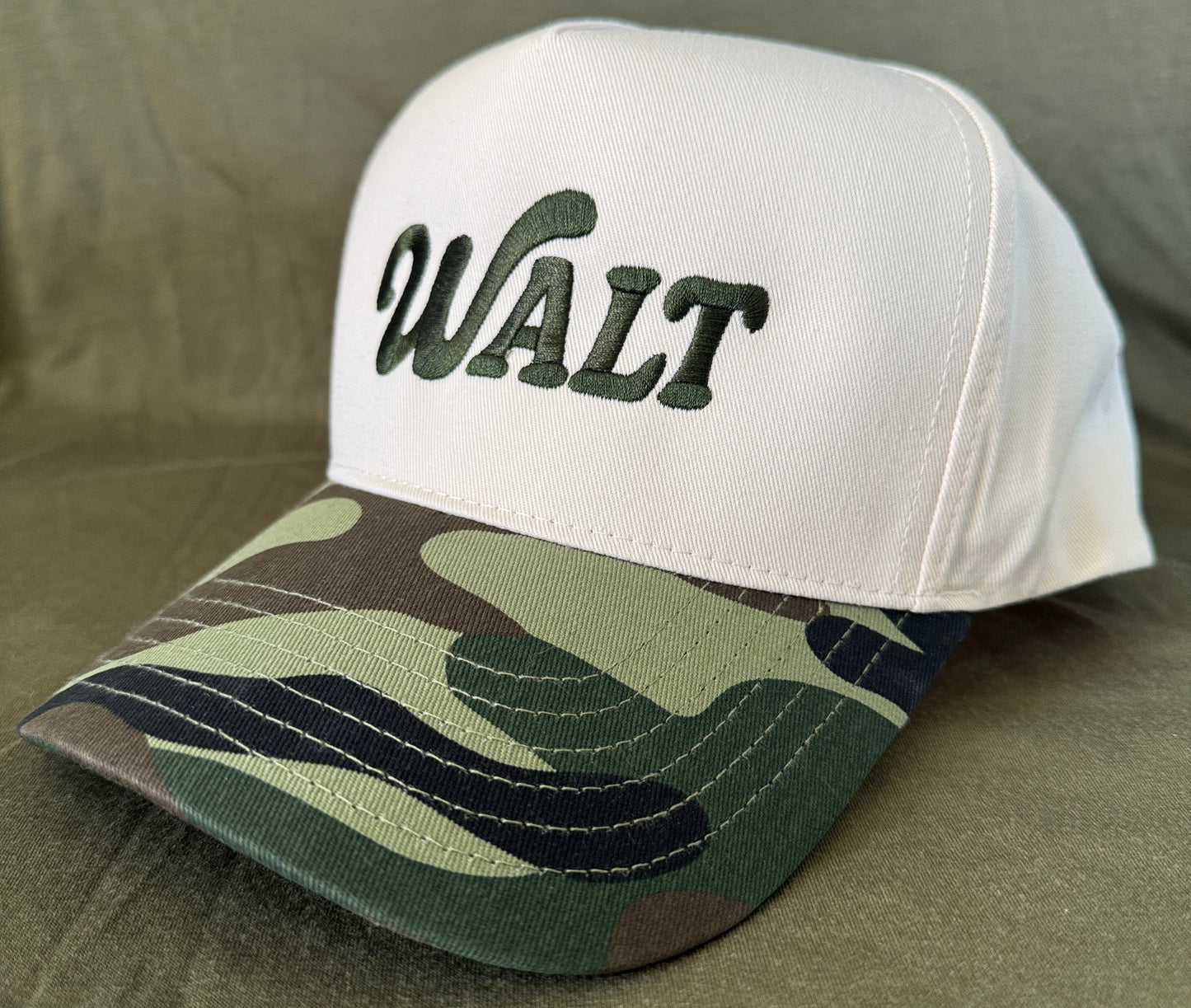 White baseball cap with green 'WALT' embroidery and camouflage bill on fabric background