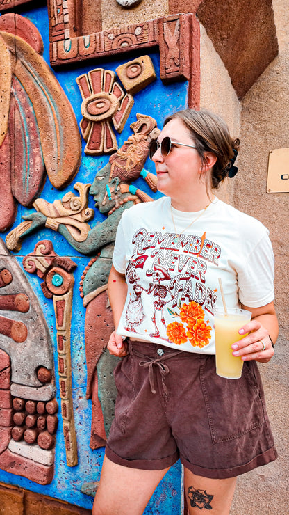Woman in sunglasses wearing a 'Remember Love Never Fades' skeleton graphic T-shirt, holding a yellow drink, standing by colorful Aztec-inspired wall art.